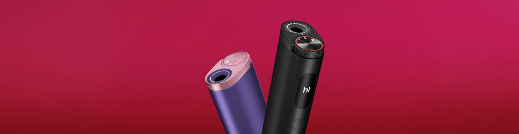 Dos elegantes aparatos electrónicos cilíndricos -uno morado con la parte superior rosa y otro negro con un toque rojo- se encuentran en posición vertical sobre un fondo rosa vibrante. El dispositivo negro muestra la palabra "hi" en su pantalla.