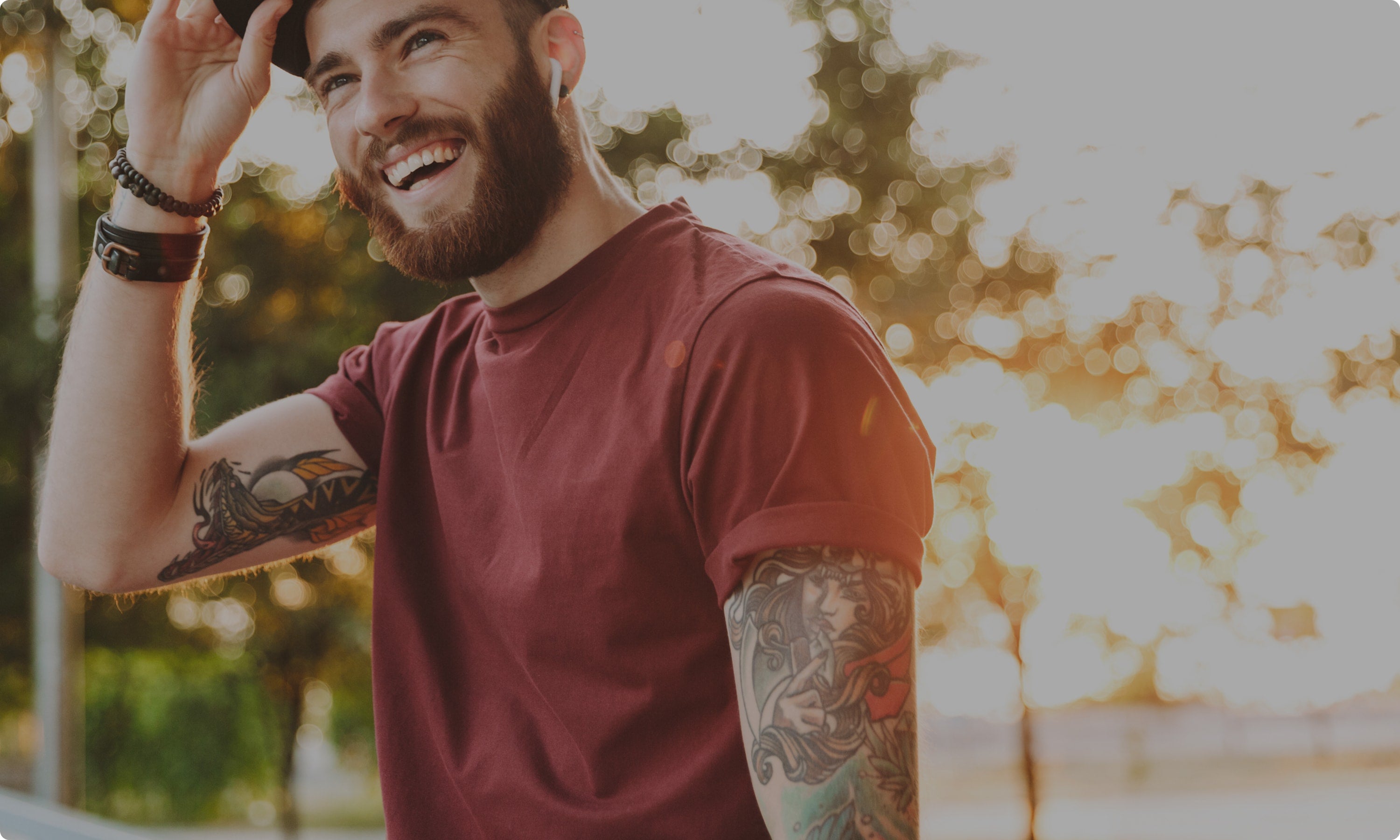 Un hombre barbudo con tatuajes, vestido con una camiseta granate y una gorra, sonríe y se levanta el ala de su sombrero al aire libre, con la luz del sol y los árboles de fondo.