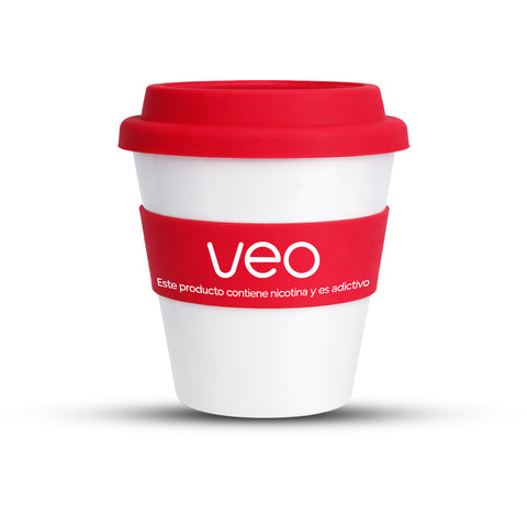 El Vaso de café Veo™ es un vaso reutilizable blanco con tapa roja y una funda en la que se lee "Veo" y "Este producto contiene nicotina y es adictivo." La taza está de pie sobre un fondo blanco. Disfruta de envío gratis con tu compra mínima.