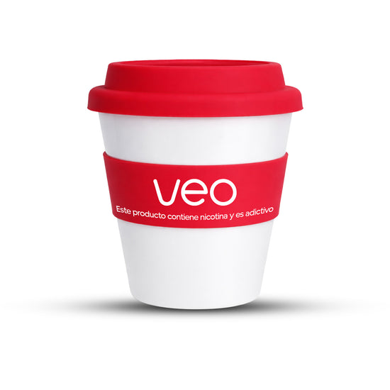 El Vaso de café Veo™ es un vaso reutilizable blanco con tapa roja y una funda en la que se lee "Veo" y "Este producto contiene nicotina y es adictivo." La taza está de pie sobre un fondo blanco. Disfruta de envío gratis con tu compra mínima.