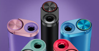 Cinco dispositivos cilíndricos de colores con mandos metálicos y la etiqueta "HEATBOOST TECHNOLOGY" están dispuestos en semicírculo sobre un fondo morado. Los dispositivos son de color negro, rosa, azul, rojo y morado.