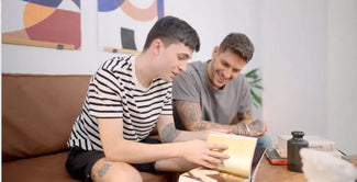 Dos hombres jóvenes se sientan en un sofá marrón, sonríen y miran juntos un libro sobre una mesa de madera. Uno lleva una camisa de rayas, el otro una camiseta gris. En la pared del fondo cuelga arte abstracto.