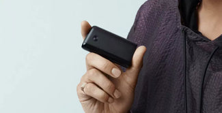 Una persona que viste una chaqueta oscura con textura sostiene un pequeño dispositivo rectangular negro entre el pulgar y los dedos sobre un fondo claro.
