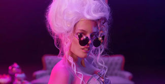 Una persona con una gran peluca blanca empolvada y gafas de sol oscuras posa bajo una dramática iluminación rosa y morada, con un top estilo corsé frente a un sofá de estilo vintage.