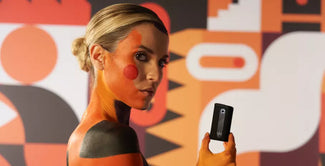 Una mujer con motivos geométricos pintados de naranja, negro y rojo en la piel sostiene un pequeño dispositivo negro. El fondo presenta atrevidas y coloridas formas abstractas en naranja, negro y blanco.