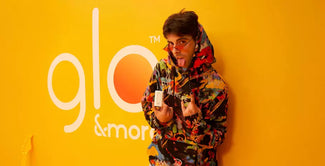 Una persona con una colorida sudadera con capucha y gafas de sol rojas posa juguetonamente delante de una pared amarilla brillante con las palabras glo & more parcialmente visibles. La persona sostiene dos dispositivos electrónicos.