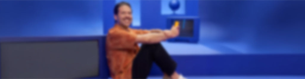 Una persona de pelo oscuro, con camisa naranja y pantalones negros, sentada en un suelo azul sobre un fondo azul, sostiene un teléfono amarillo y sonríe. La imagen está muy borrosa.