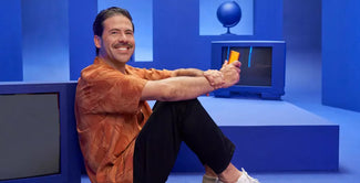Una persona de pelo oscuro y bigote, con una camisa naranja estampada, pantalones negros y zapatillas de deporte, se sienta sonriente en el suelo de una habitación azul con pantallas de televisión y decoración geométrica, sosteniendo un polo naranja.
