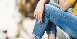 Una persona vestida con unos vaqueros azules arremangados y una camisa amarilla está sentada en unos escalones exteriores, sosteniendo un cigarrillo entre los dedos. La escena es informal y no se ve el rostro de la persona.