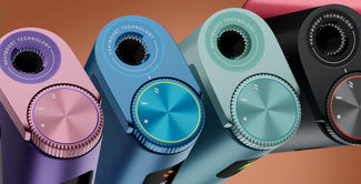 Primer plano de cuatro dispositivos electrónicos de color púrpura, azul, verde azulado y negro, cada uno con un gran dial circular con la etiqueta HeatBoost Technology y mandos de control texturizados, dispuestos uno al lado del otro sobre un fondo marrón.