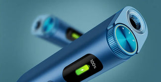 Primer plano de un aparato de aseo eléctrico azul con una pantalla digital que muestra el 100% de la batería y un símbolo verde de carga. Otro aparato similar aparece borroso en el fondo.